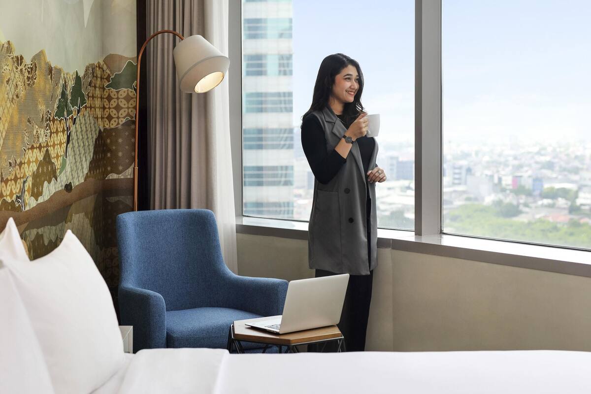 A hotel room with a large window, a blue armchair, a desk with a laptop, and a person holding a mug.