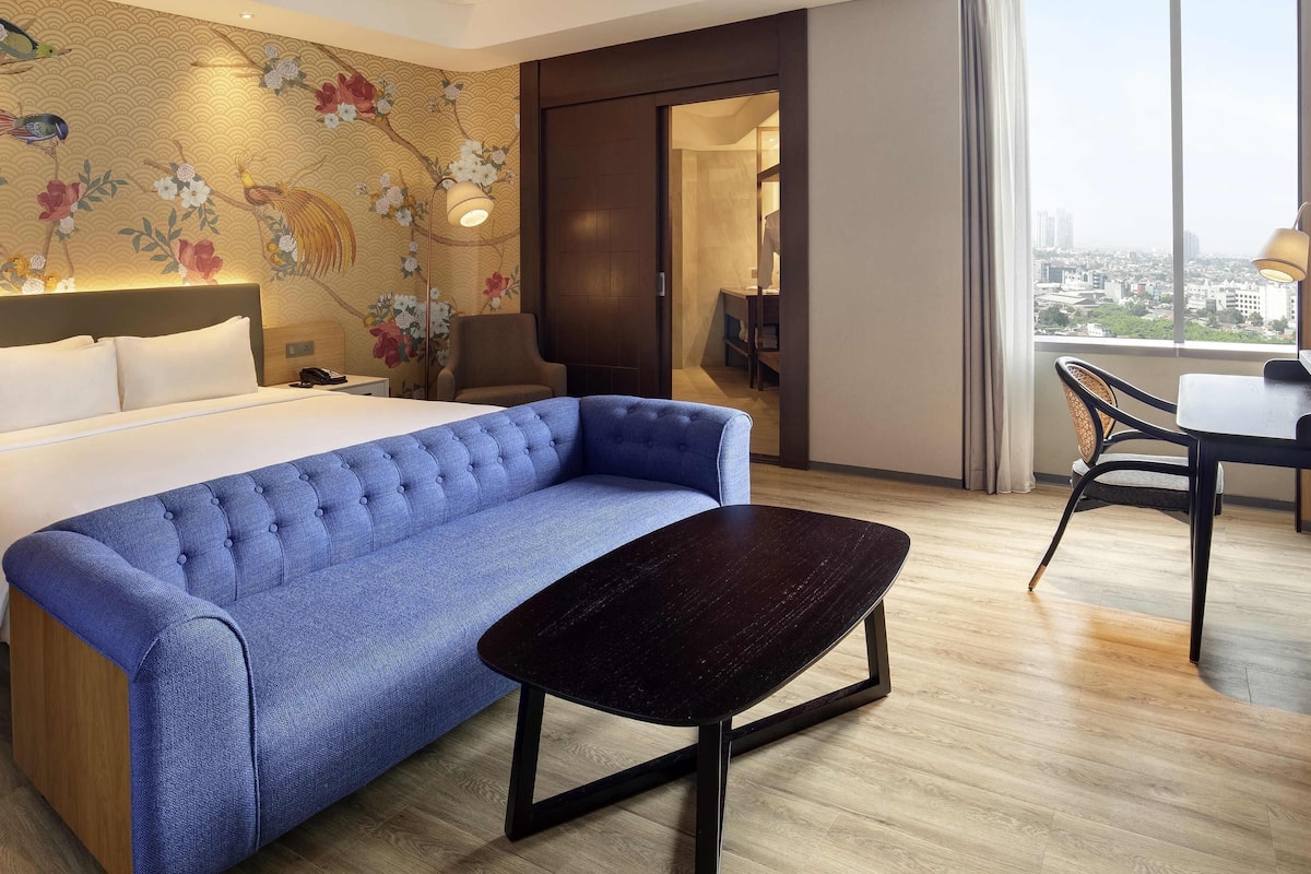 Ein Hotelzimmer mit einem Bett, einem blauen Sofa, einem kleinen Tisch, einem Stuhl, einer Wand mit floralem Wandpapier und einem großen Fenster mit Blick auf die Stadt.