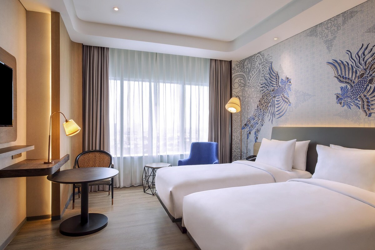 A hotel room with two beds, a desk, a chair, a TV, and a large window with curtains.