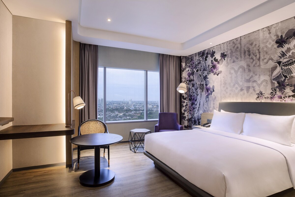 A hotel room with a bed, a desk, a chair, a small table, and a view of the city.