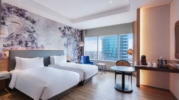 A modern hotel room with two beds, a small dining table, a chair, a desk with a lamp, and a large window offering a city view.