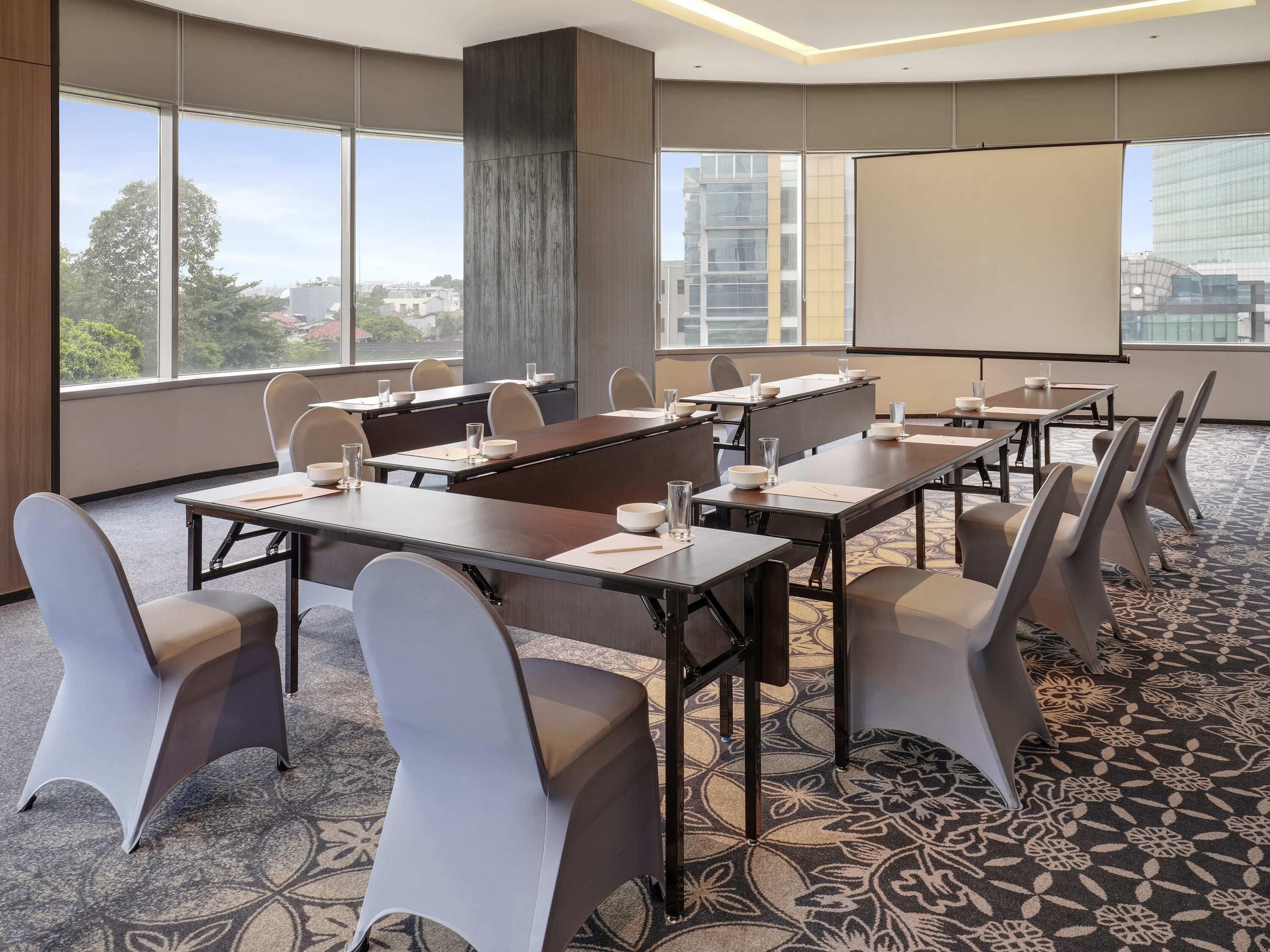Sala de conferências com mesas, cadeiras, tela de projetor e amplas janelas com vista para a cidade.
