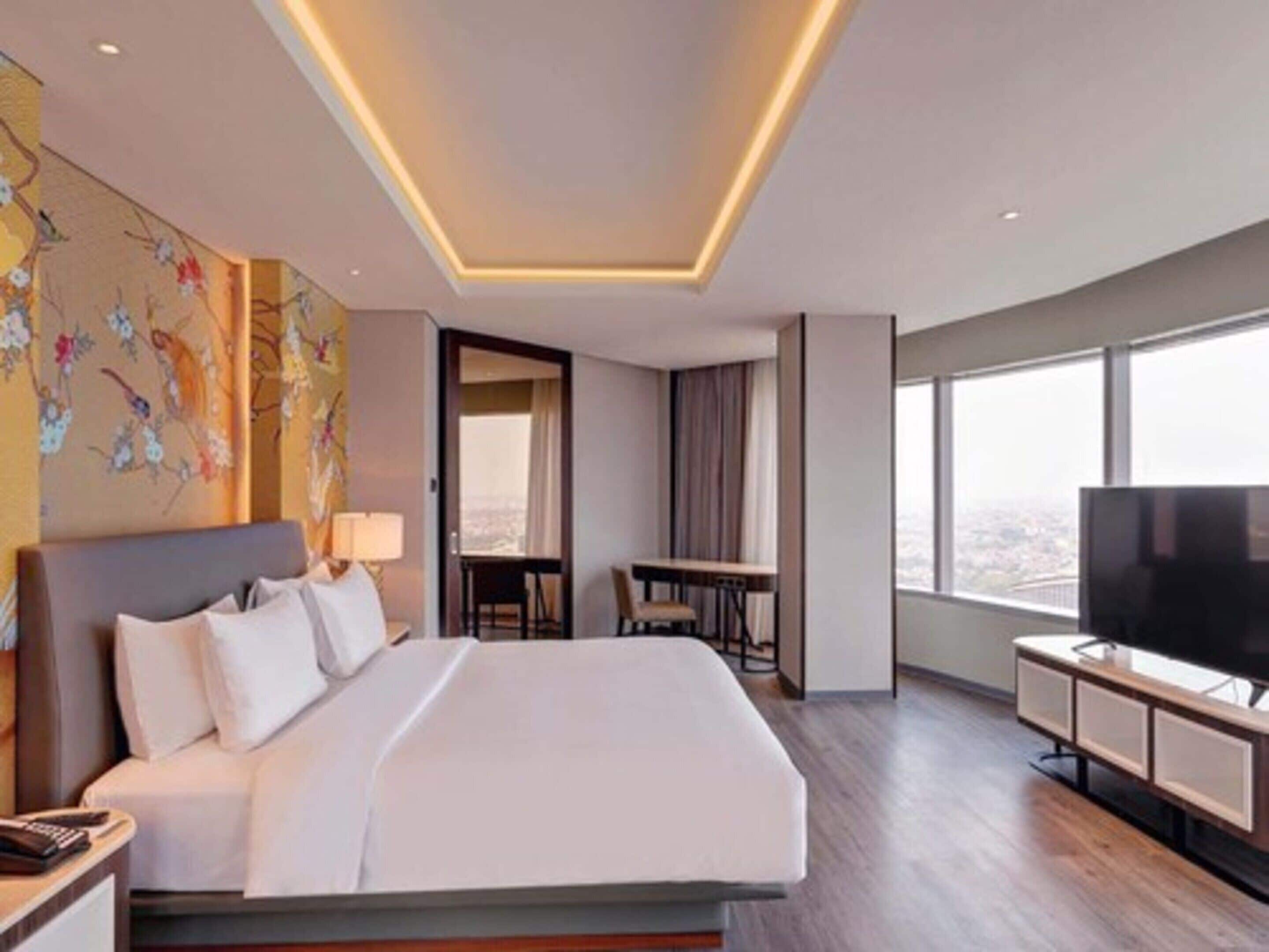 A modern hotel room with a large bed, a desk, a television, and a city view.