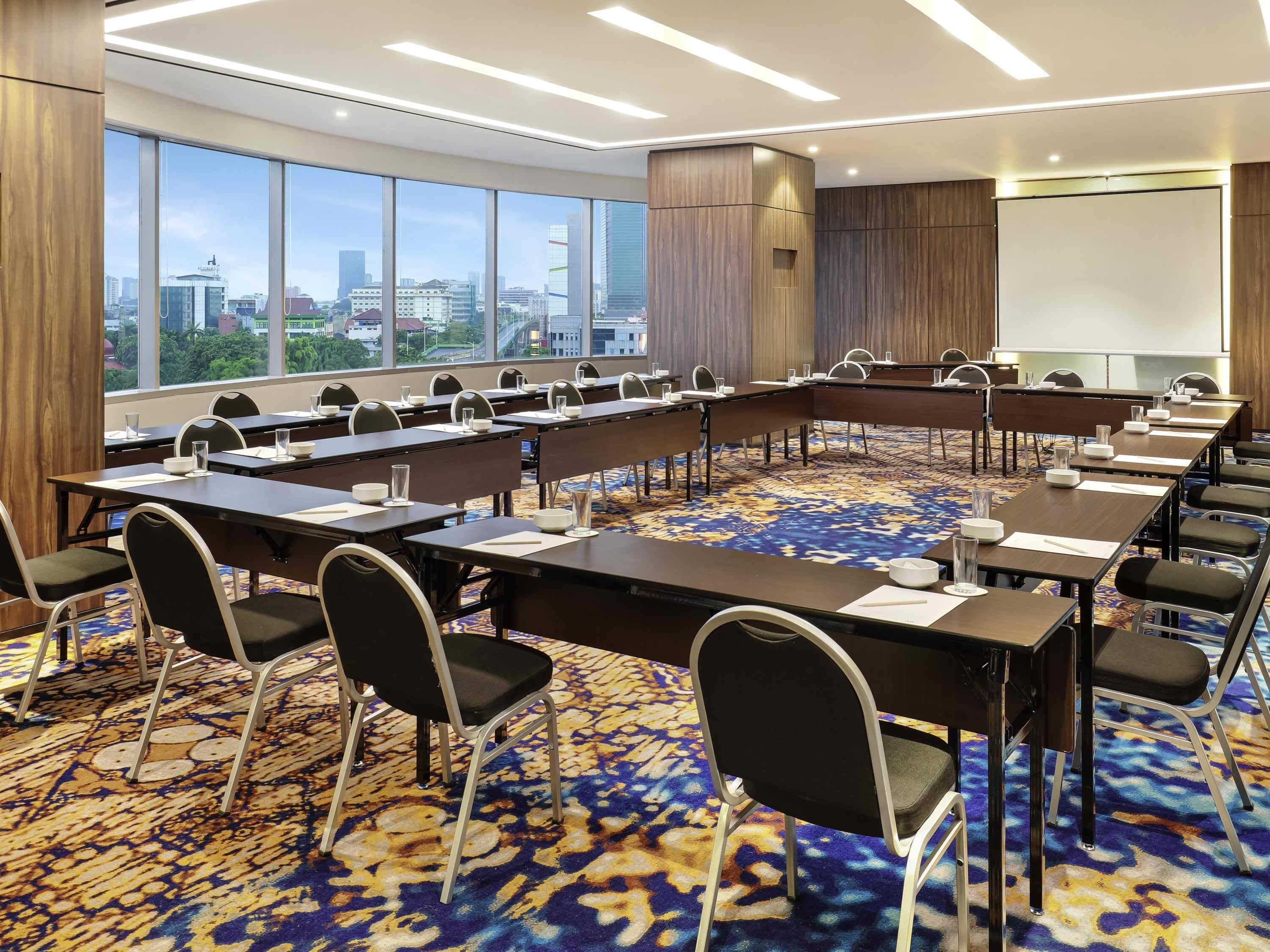 Een congreszaal met lange tafels, stoelen, een projectiescherm en grote ramen met uitzicht op de stad.