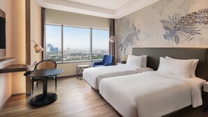 Habitación de hotel con dos camas, un escritorio, una silla, una mesita y un amplio ventanal con vistas a la ciudad.