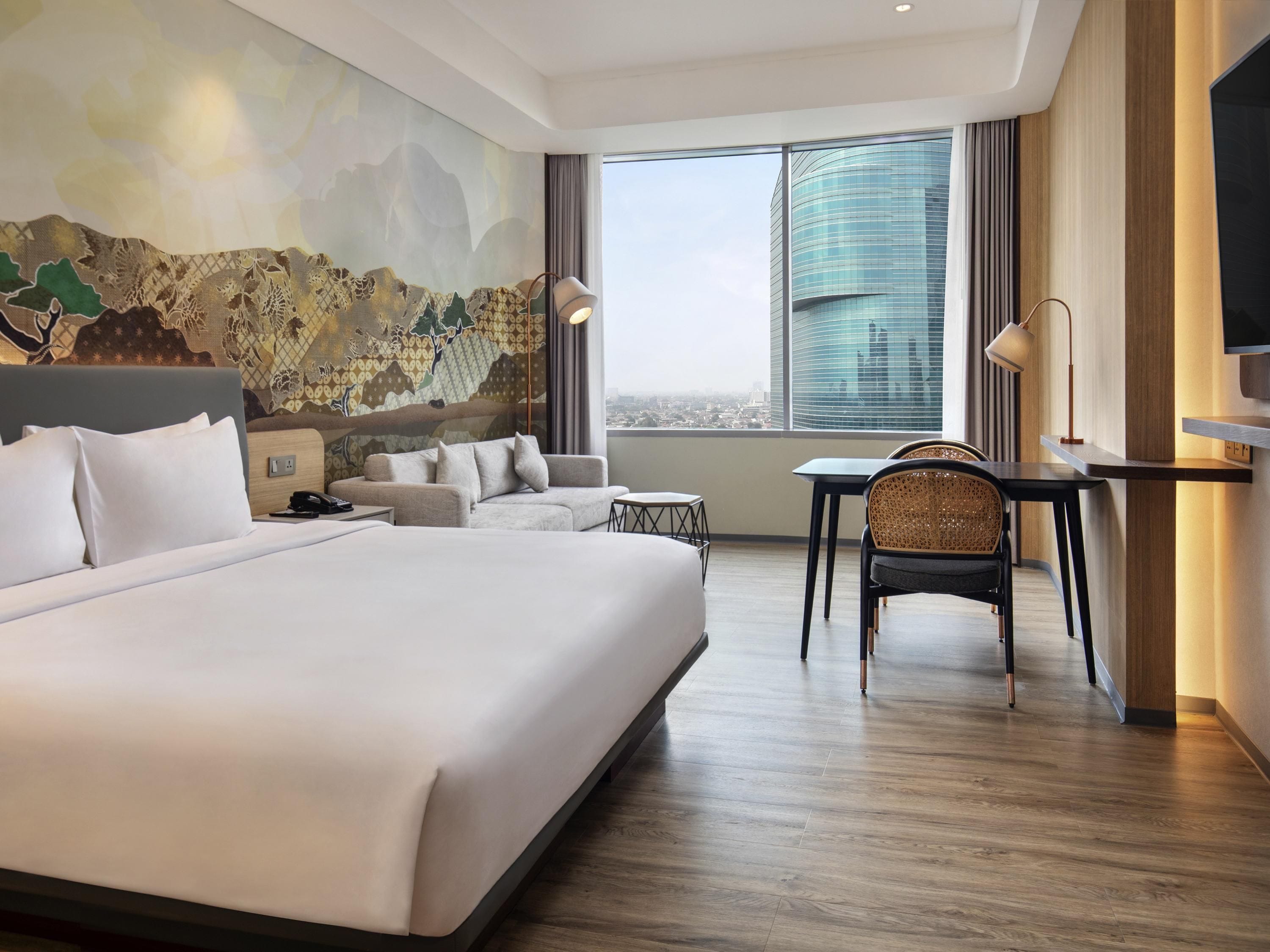 A hotel room with a large bed, a sofa, a desk, and a chair. There is a cityscape view through the window.