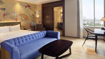 Ein Hotelzimmer mit einem Bett, einem blauen Sofa, einem kleinen Tisch, einem Stuhl, einer Wand mit floralem Wandpapier und einem großen Fenster mit Blick auf die Stadt.