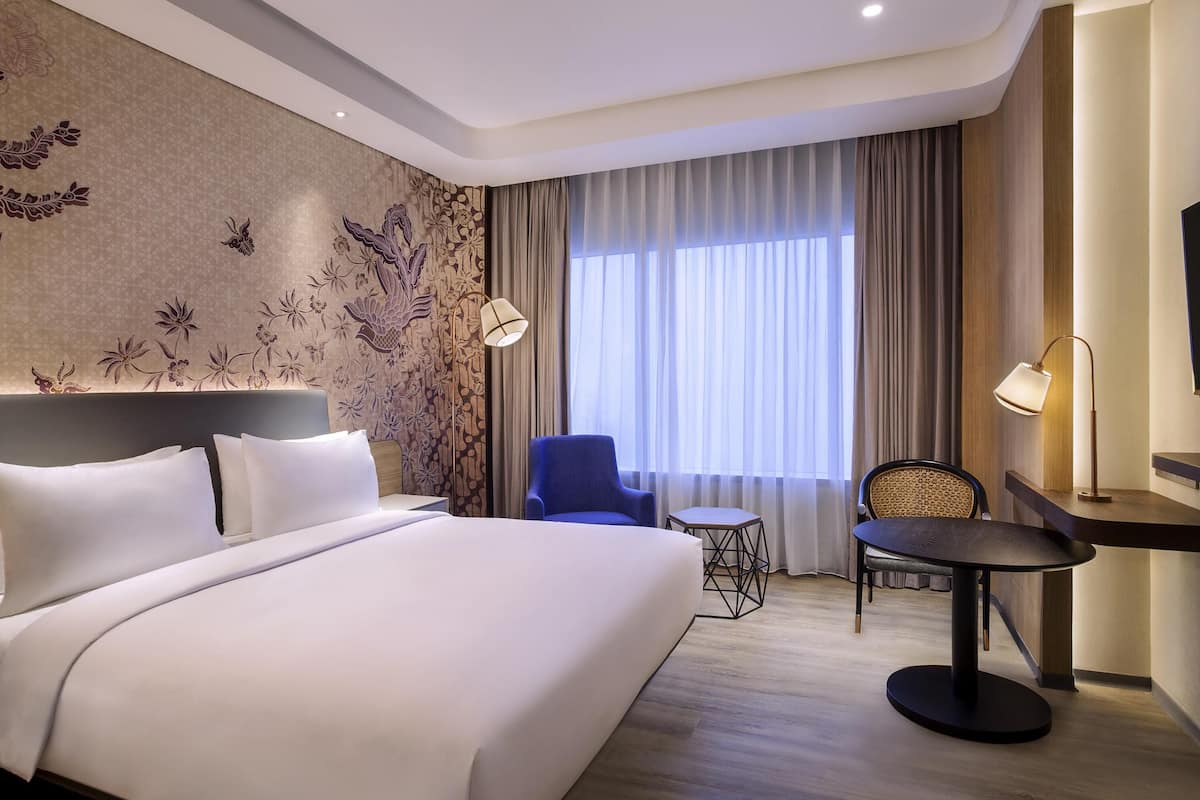 Ein Hotelzimmer mit einem großen Bett, einem Schreibtisch, zwei Stühlen, einem kleinen Tisch und einem großen Fenster mit Vorhängen.
