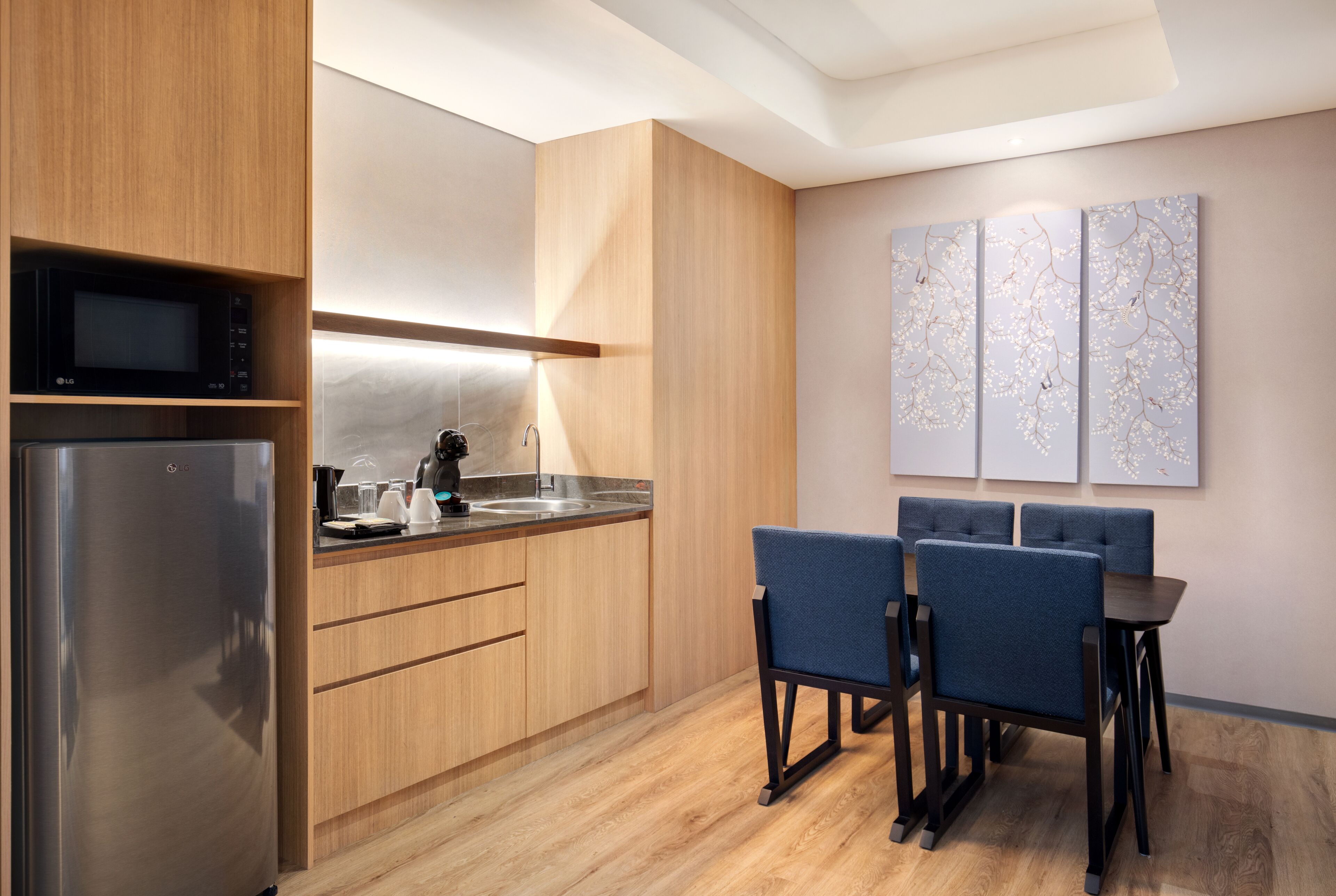 A modern kitchen with wooden cabinets, a stainless steel refrigerator, a microwave, and a coffee maker. There is a dining area with a table and chairs.