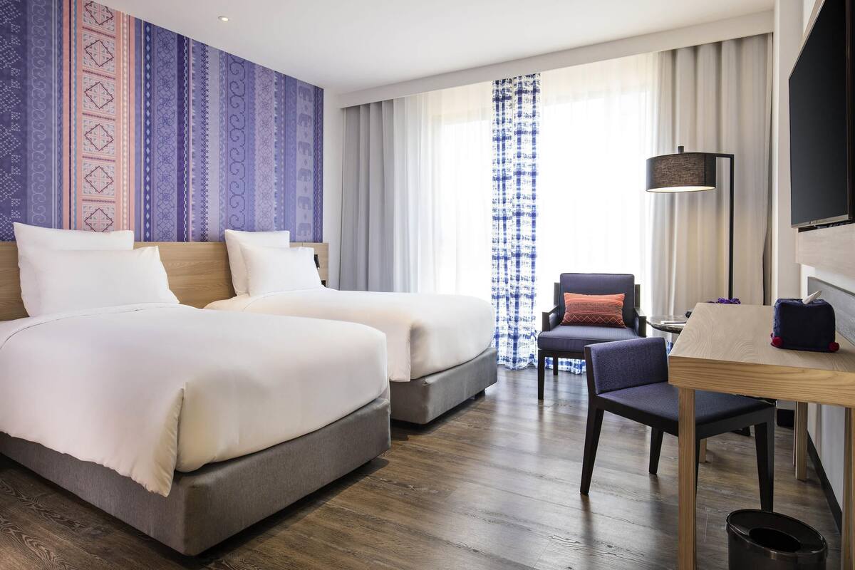 Une chambre d’hôtel avec deux lits, un bureau, une chaise, une télévision et une grande fenêtre avec des rideaux.