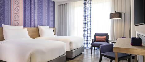 Habitación de hotel con dos camas, un escritorio, una silla, un televisor y un ventanal con cortinas.