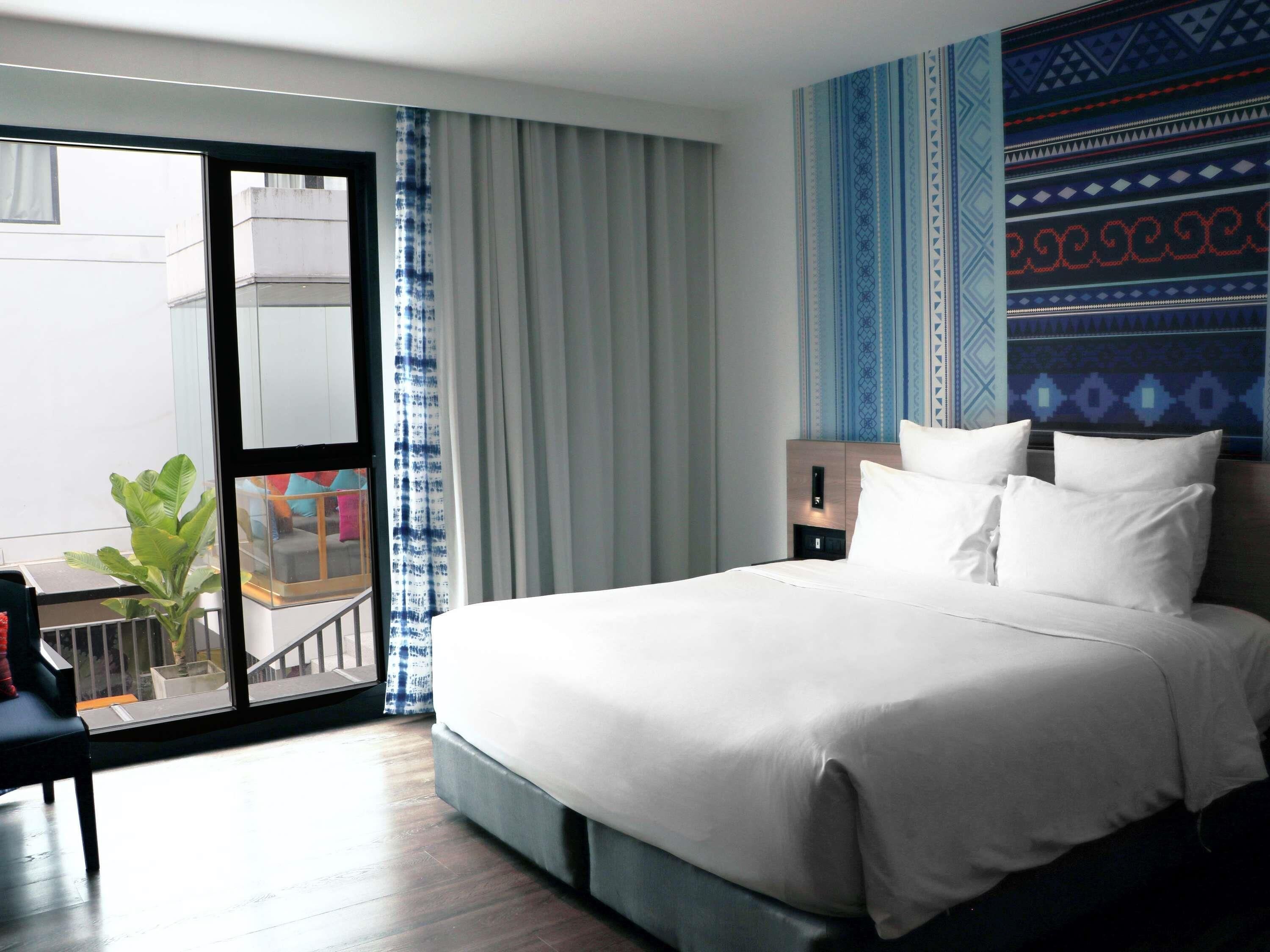 A hotel room with a large bed, a nightstand, a desk with a chair, and a view through a sliding glass door to an outdoor area.