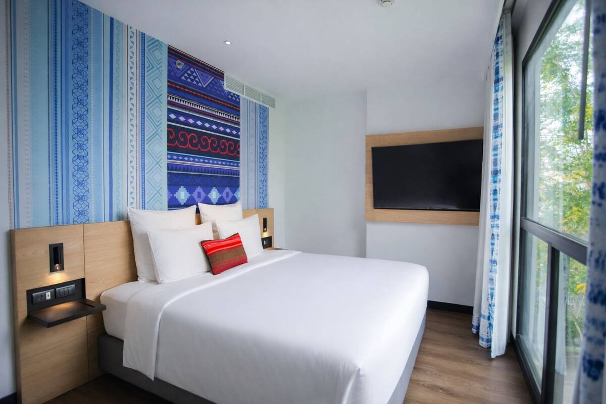 Une chambre d’hôtel avec un grand lit, une table de chevet avec une lampe, un téléviseur fixé au mur et une fenêtre avec des rideaux.