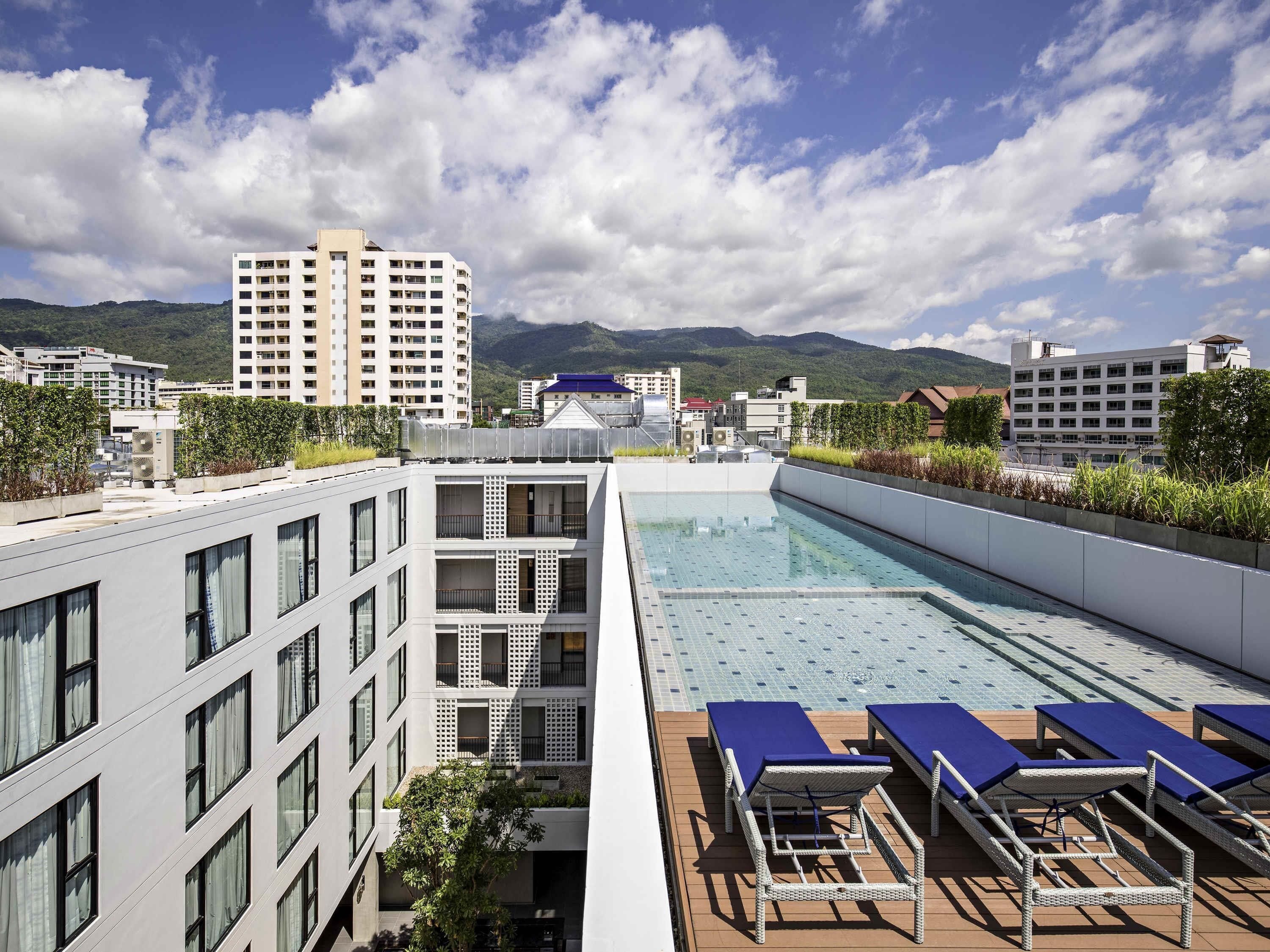 A rooftop swimming pool with lounge chairs, a view of surrounding buildings, and a mountainous backdrop.