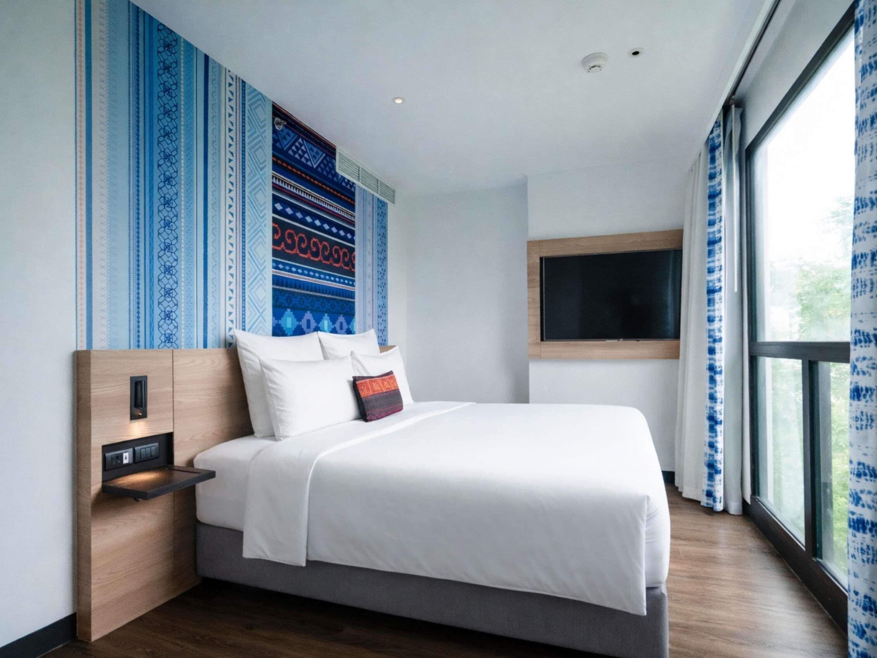 A modern hotel room with a large bed, a built-in TV, a bedside table with a power outlet, and a window with curtains.