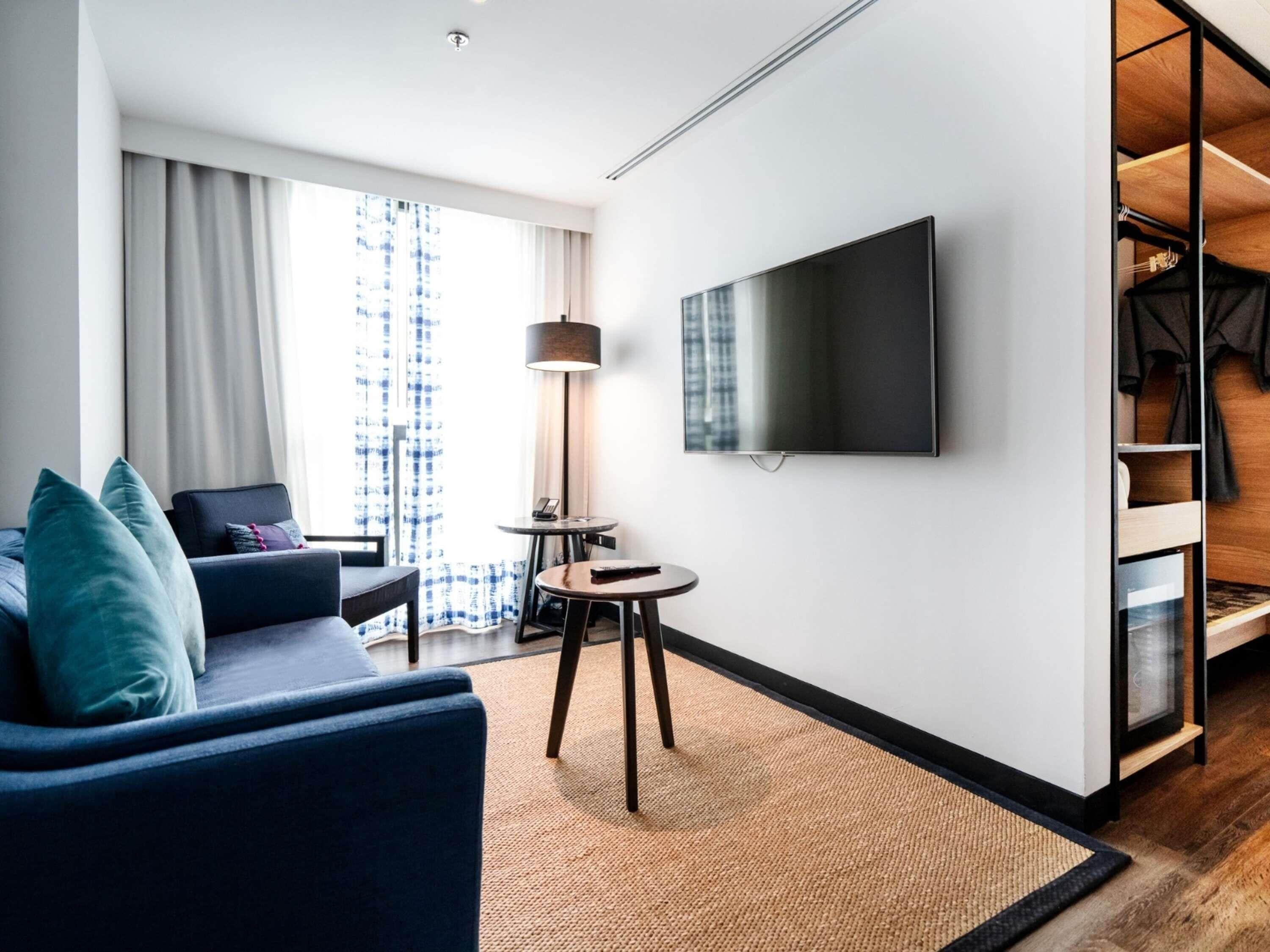 Habitació d'hotel moderna amb un sofà, una televisió de pantalla plana, una làmpada i vista a la ciutat a través de grans finestres.