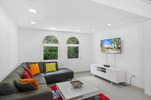 A modern living room with a grey sofa, colorful cushions, a white coffee table, and a flat-screen TV mounted on the wall. - Spacious 4BR Home w/ Private Pool & Backyard (Noord)