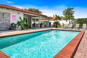 A swimming pool with a tiled edge, a covered patio area with chairs, and a building with a tiled roof.