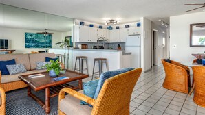 A modern living room with a sofa, coffee table, and wicker chairs. A kitchen with white cabinets and blue accents is visible in the background.