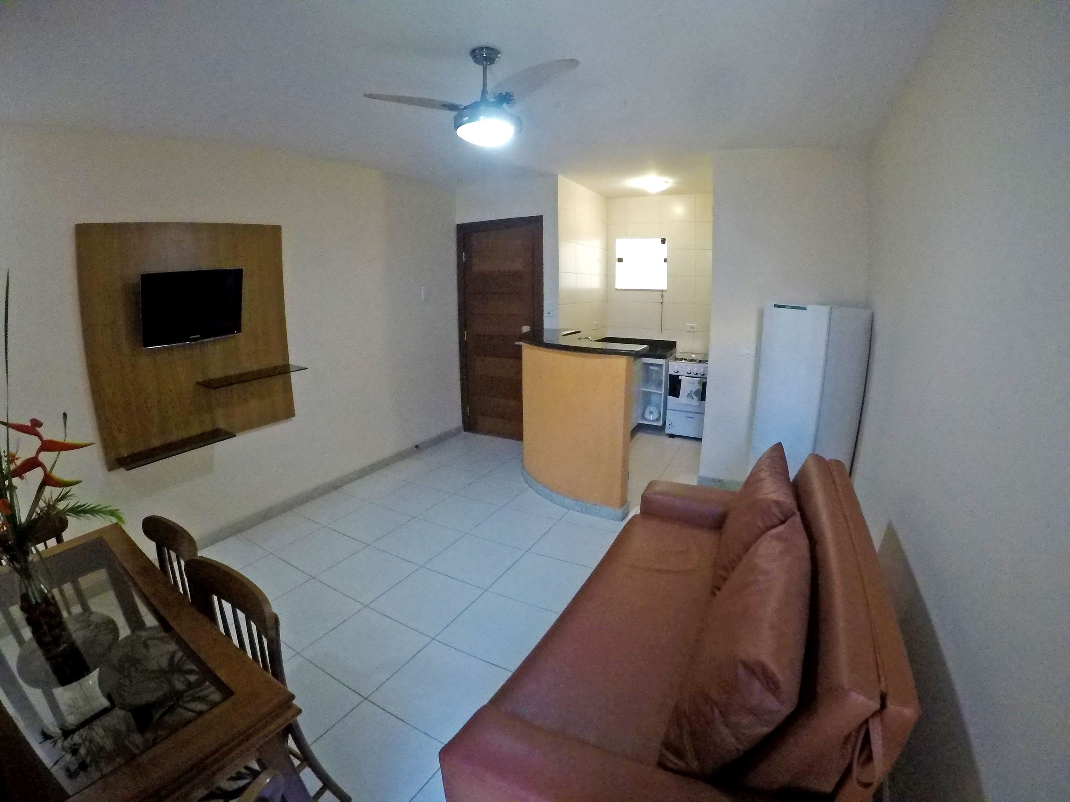 A living room with a sofa, a TV mounted on the wall, a ceiling fan, and a kitchen area with a stove and refrigerator.