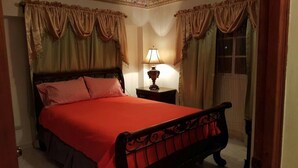 A neatly made bed with a red bedspread and checkered pillows, a bedside table with a lamp, and curtains with a floral pattern. - Apartamento Florangel (Santo Domingo Este)