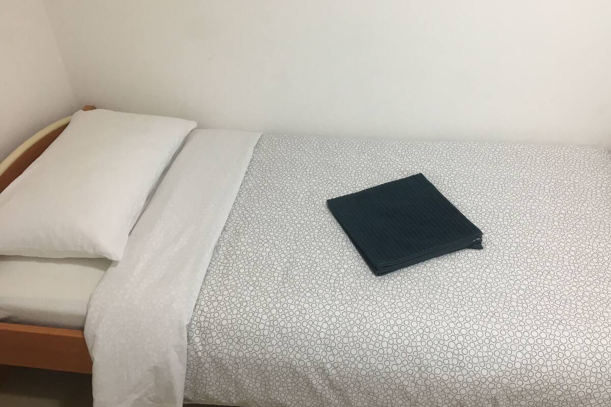 Une petite chambre avec un lit simple, recouverte d’une courtepointe blanche et grise à motifs, un oreiller blanc et une couverture foncée pliée.