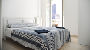 Una cama bien hecha con ropa de cama estampada, un cabecero blanco y vistas a un paisaje urbano a través de una puerta abierta.