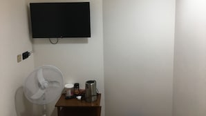 Una habitación con una televisión de pantalla plana montada en la pared, una pequeña mesa de madera con un ventilador, una cafetera y un control remoto.