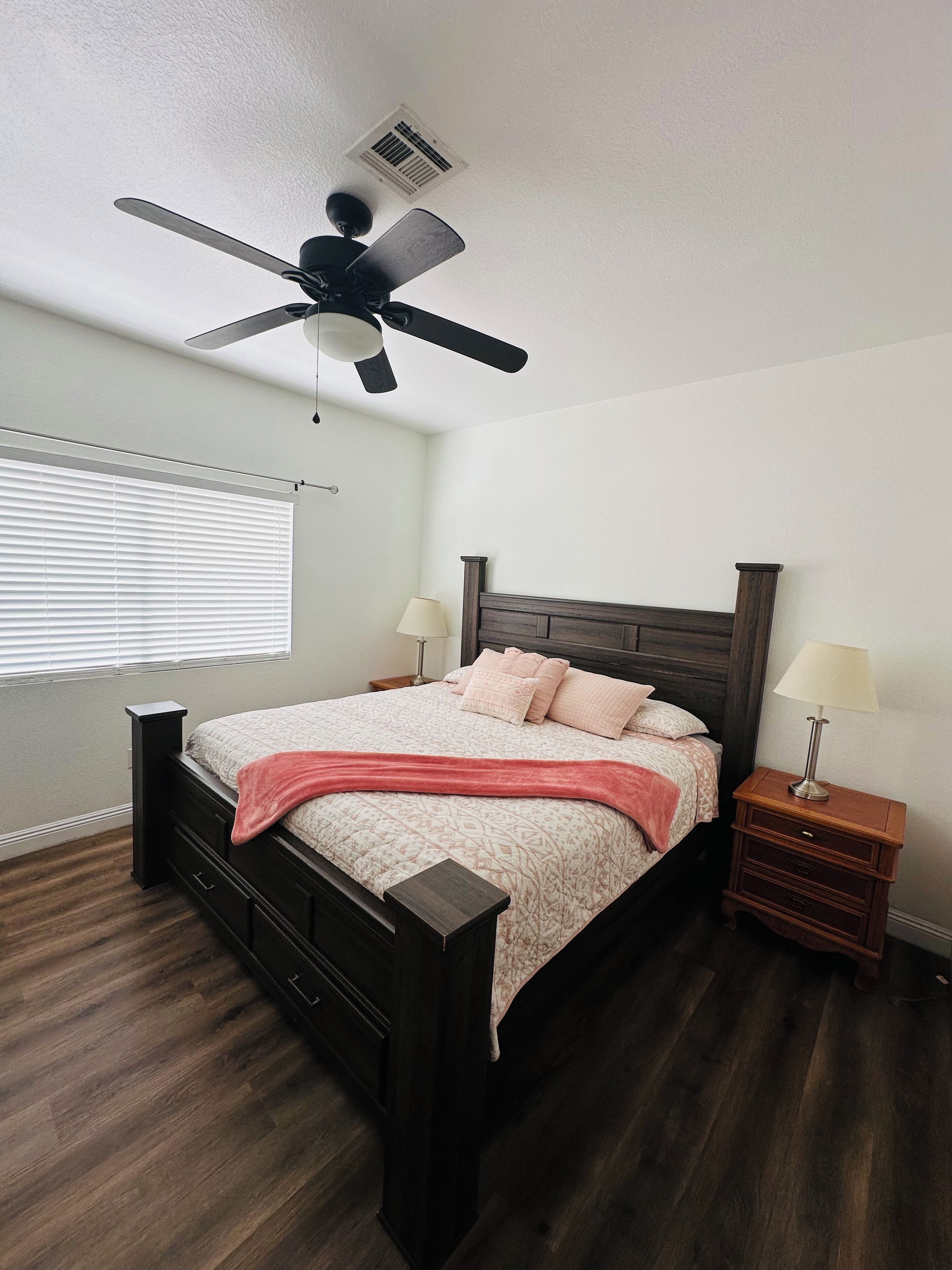 A bedroom with a bed, a ceiling fan, a nightstand with a lamp, and a window with blinds.