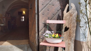 Une porte en bois rustique avec un loquet en métal, un banc en bois orné d’une découpe en forme de cœur, et un pot de fleurs posé sur le banc.
