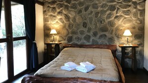 A bedroom with a stone wall, a wooden bed frame, two bedside tables with lamps, and a window with curtains. - Cabanas Merlin (Merlo)