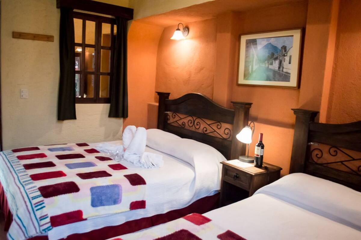 A hotel room with two beds, a nightstand with a lamp and a bottle, a window with curtains, and a painting on the wall.