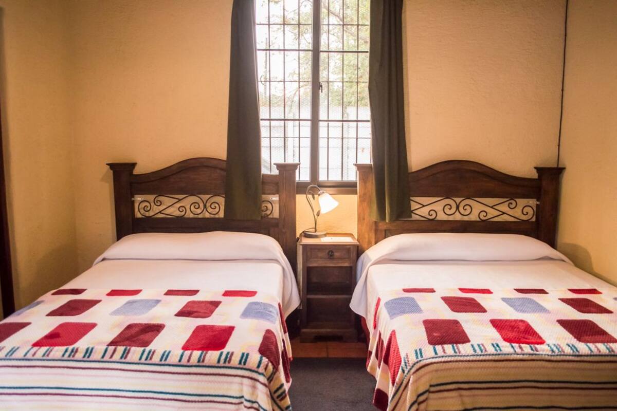 A room with two beds, each with a patterned bedspread and a wooden headboard. There is a nightstand with a lamp between the beds.