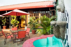 An outdoor patio with red umbrellas, a stone fountain, and potted plants. - Hotel Palma De Mallorca (Antigua Guatemala)