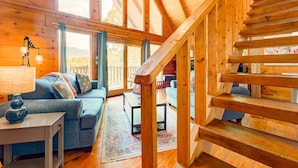 Un accogliente interno di una baita con una scala in legno, un divano, un tavolino da caffè e ampie finestre.