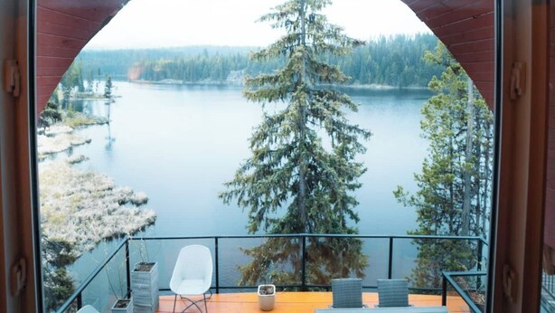 Een houten terras met twee witte stoelen en een kleine tafel, met uitzicht op een meer met bomen en een verre oever.