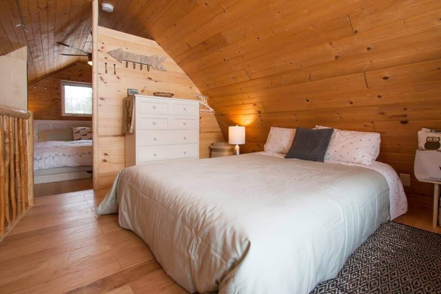 Une chambre mansardée chaleureuse avec un plafond en bois, un lit blanc, une commode et une petite table avec une lampe.