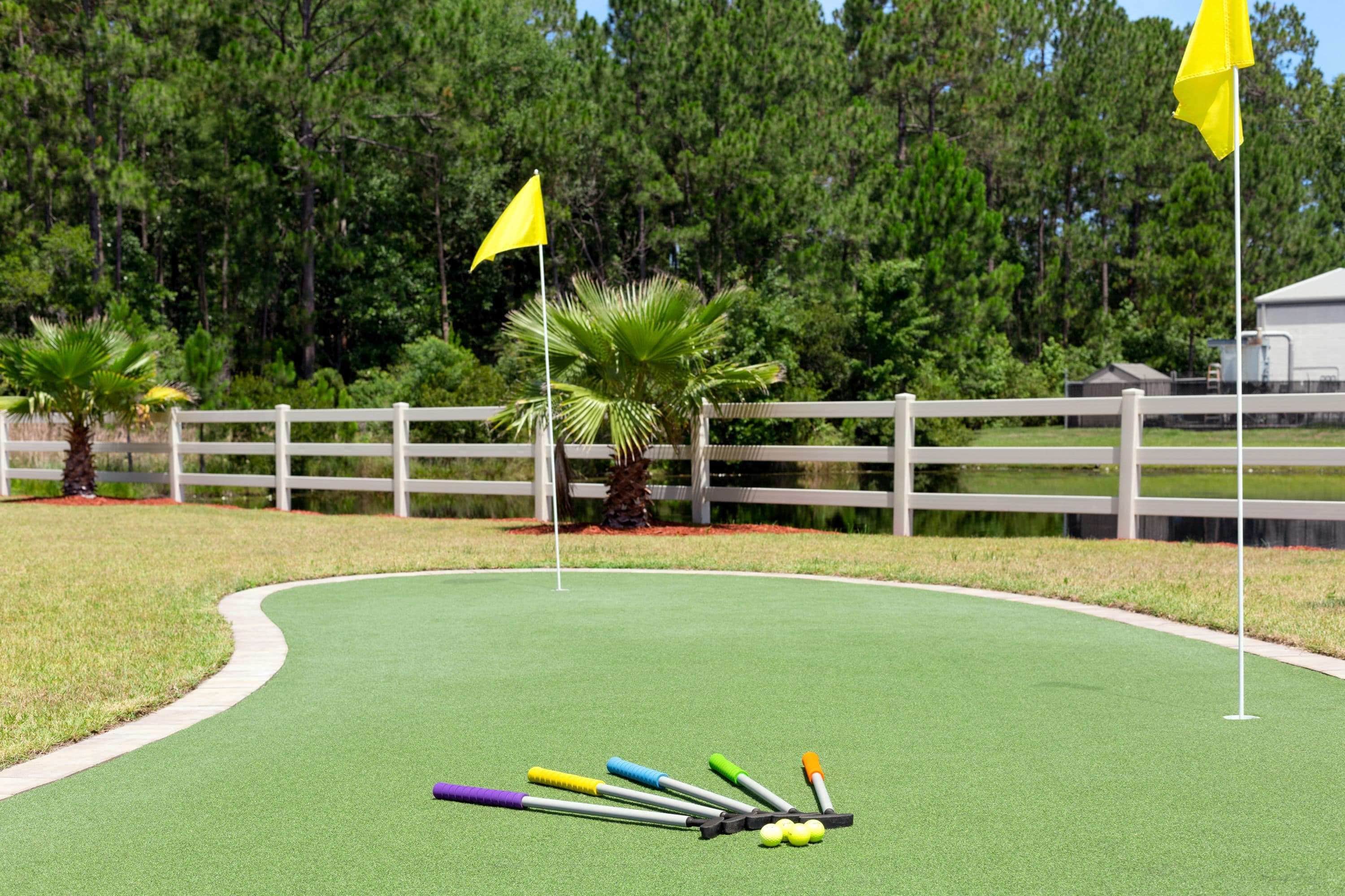 Un campo de golf con césped artificial verde, una valla blanca, palmeras y banderas amarillas.