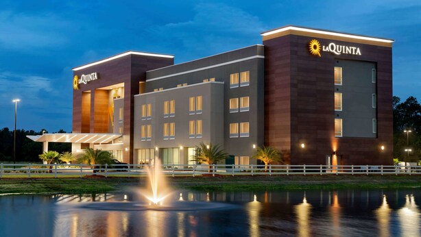 Ein modernes Hotel mit einem gut sichtbaren "La Quinta"-Schild, einem gut beleuchteten Eingang und einer reflektierenden Wasserspiegelung im Vordergrund.
