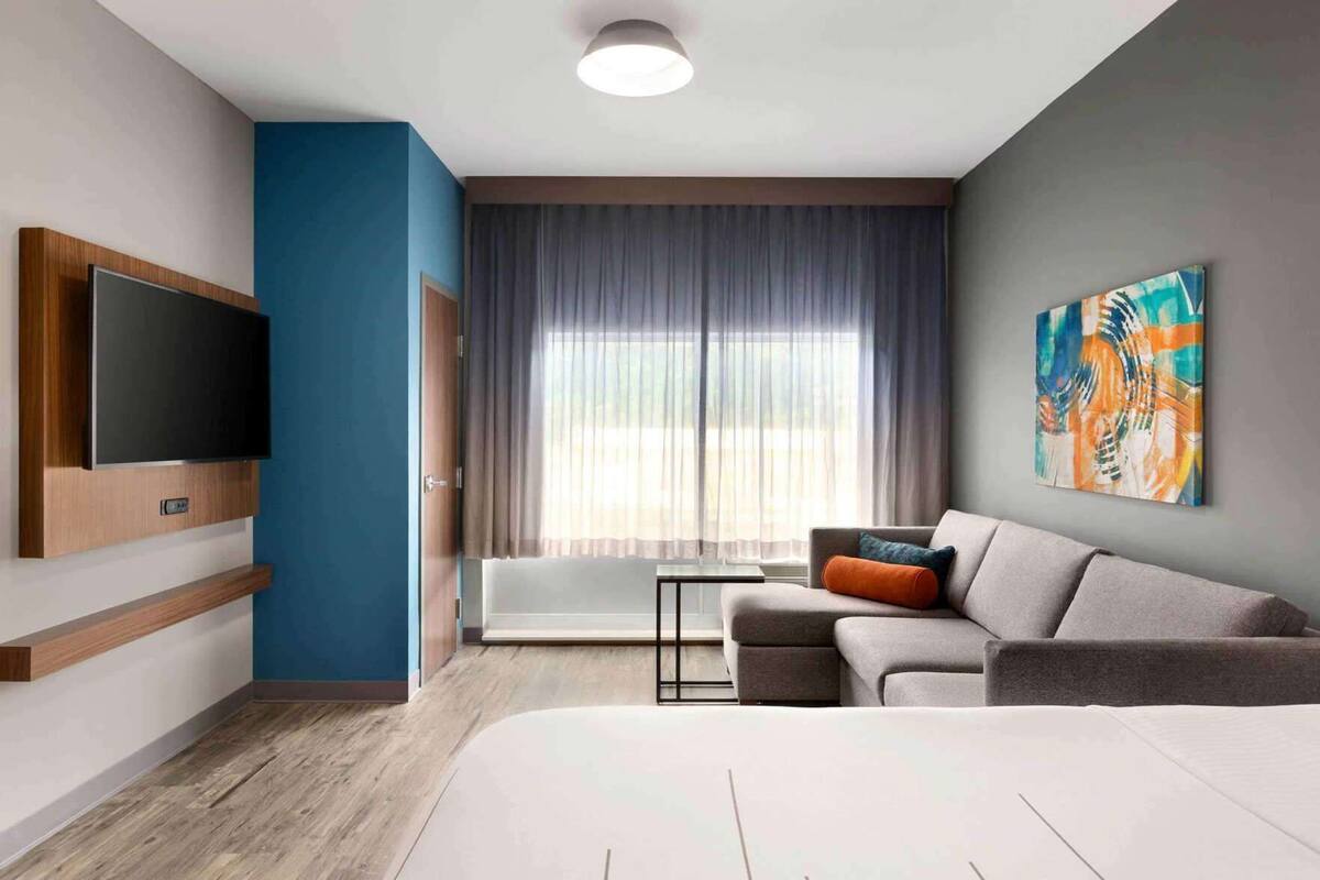A modern hotel room with a flat-screen TV, a sofa with cushions, a large window with curtains, and a painting on the wall.