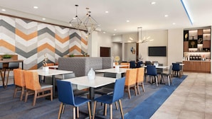 Eine moderne Hotelhalle mit Wandgestaltung im geometrischen Muster, verschiedenen Sitzbereichen mit blauen und orangefarbenen Sesseln und einer Bar mit Holzausstattung.