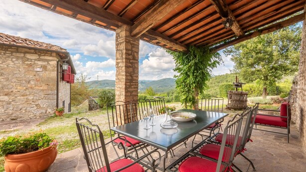 Ein überdachter Essbereich im Freien mit Tisch und Stühlen, ein Steinhaus und Blick auf die Landschaft.
