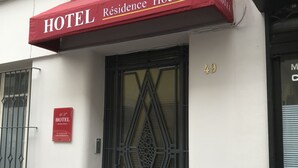 Eine Hotelfassade mit einer roten Markise mit der Aufschrift „HOTEL Residence Hoche“, einem schwarzen Metallgitter und einem Fenster mit dekorativem Gitter.