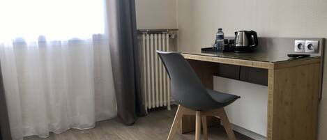 Una habitación moderna con un escritorio de madera, una silla negra, un radiador, una ventana con cortinas y un hervidor.