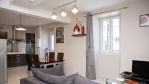 Ein modernes Wohnzimmer mit grauem Sofa, einem Essbereich mit Tisch und Stühlen, einer Küche mit Herd und Schränken sowie einem Fernseher auf einem Schrank.