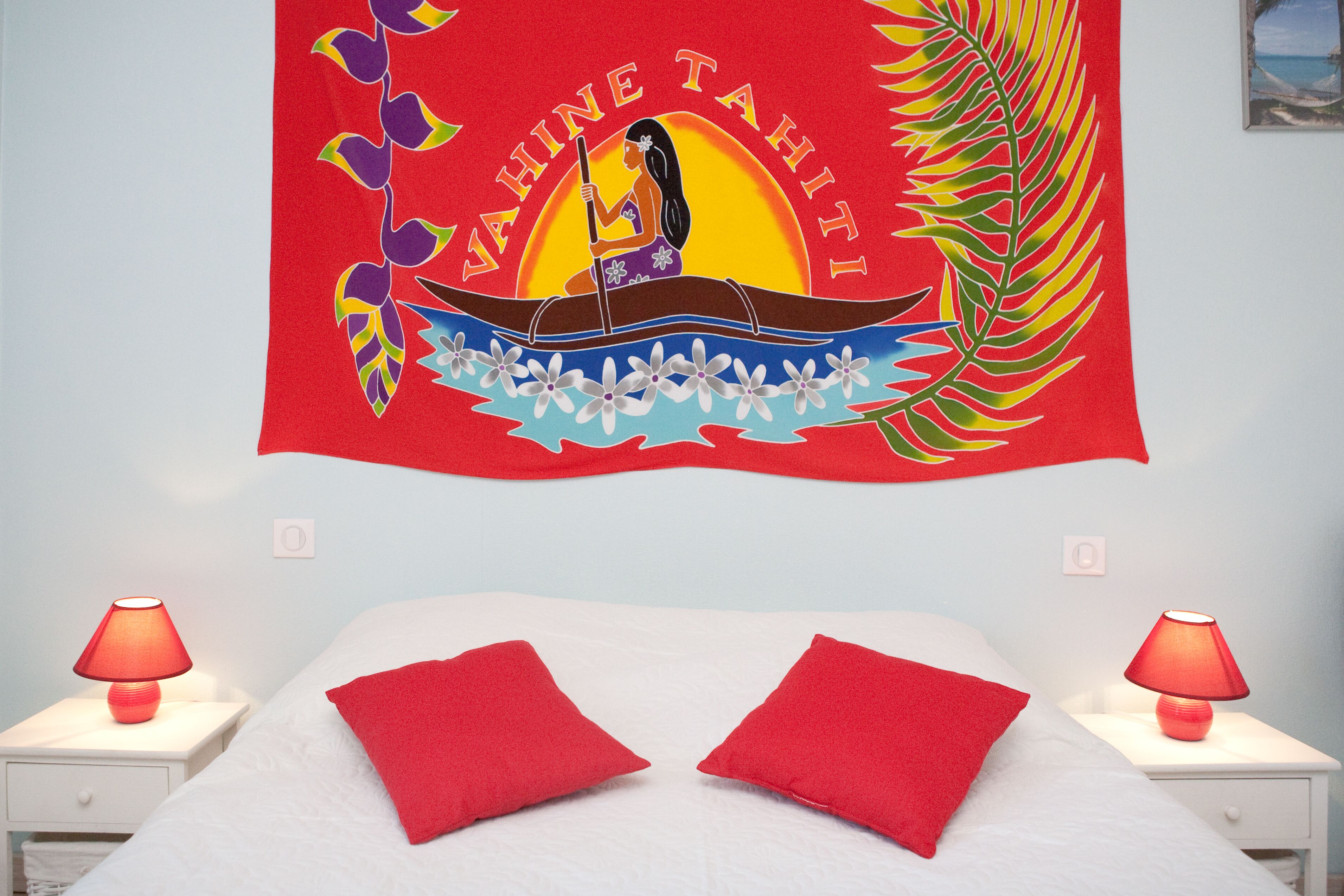 A bed with red pillows, a wall tapestry with a tropical design, and two bedside lamps.