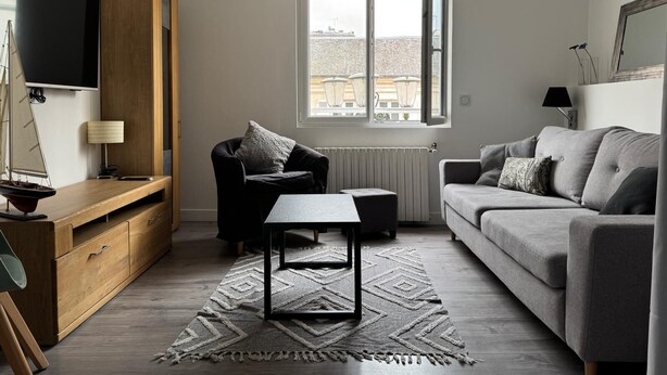 A modern living room with a sofa, a chair, a small table, a TV, and a window with a view of buildings.