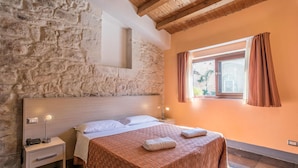 Quarto com parede de pedra, teto de madeira, cama com colcha estampada, duas luminárias de cabeceira e uma janela com cortinas.