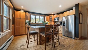 A modern kitchen with wooden cabinets, a central island with bar stools, stainless steel appliances, and large windows offering views of trees.