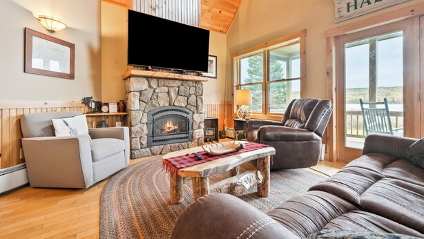 A cozy living room with a stone fireplace, a flat-screen TV mounted above it, a comfortable sofa, a leather armchair, and a large window offering a view of the outdoors.